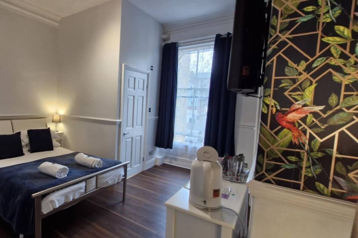 A hotel room with a bed, bedside tables, a desk with a chair, a window with curtains, and a wall with a geometric pattern.