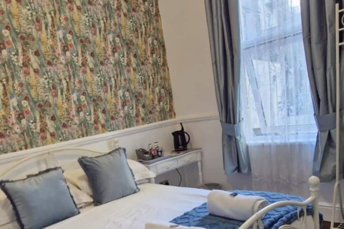 A bedroom with a bed, a nightstand, a window with curtains, and a floral tapestry on the wall.