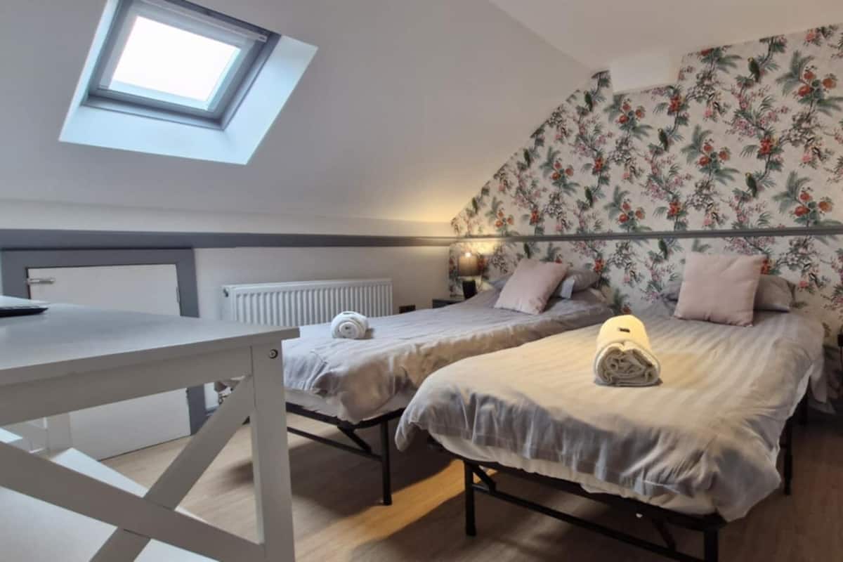 A bedroom with two beds, a skylight, floral wallpaper, bedside lamps, and a wooden floor.