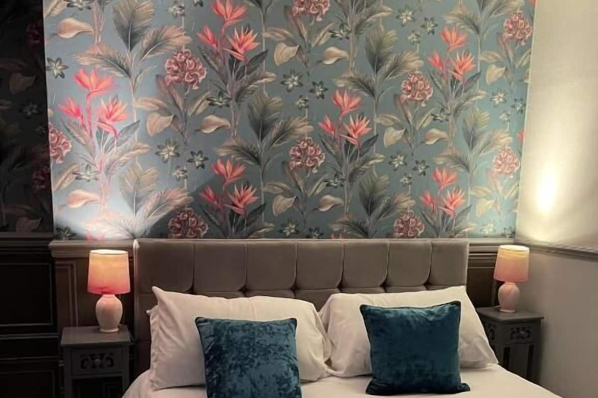 A bedroom with a floral wallpaper, a tufted headboard, two bedside lamps, and a blue bedspread.