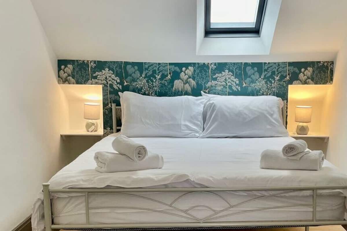 A neatly made bed with two pillows and two rolled towels, a skylight above, and floral wallpaper on the walls.