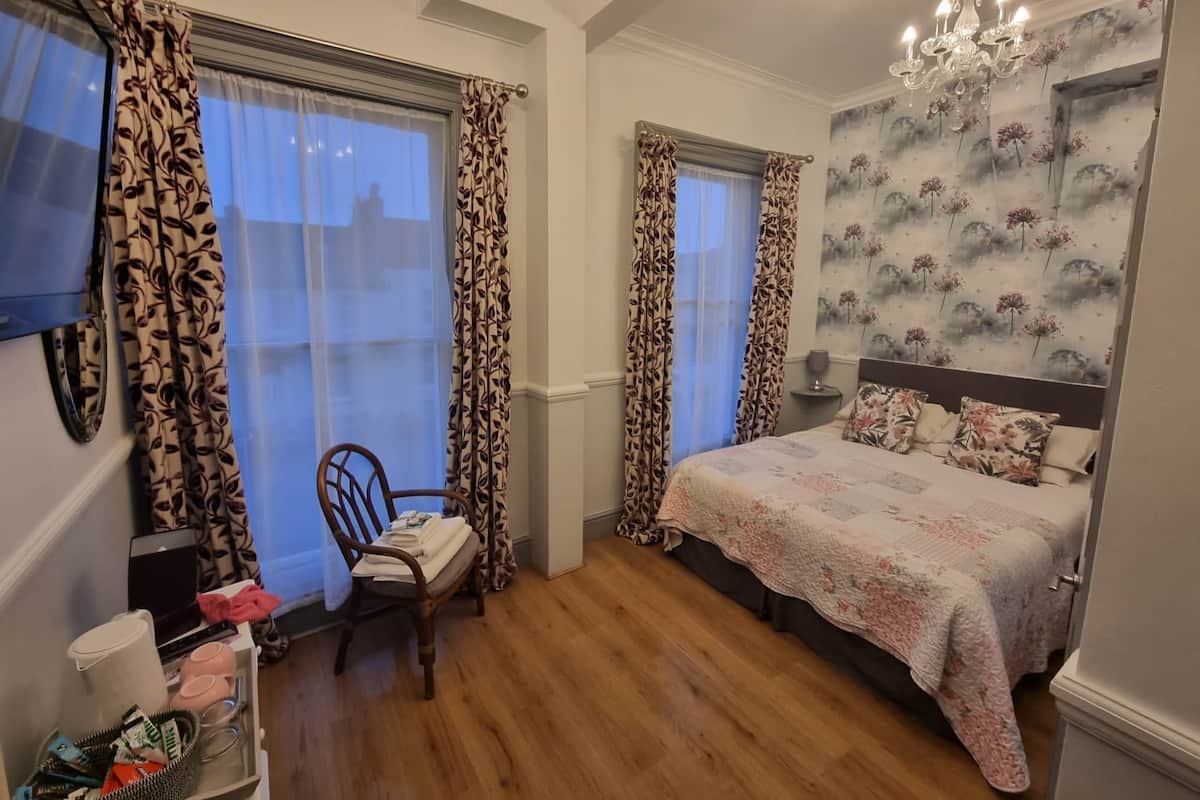 A bedroom with a bed, two windows with patterned curtains, a wooden chair, a desk with a laptop, and a chandelier.