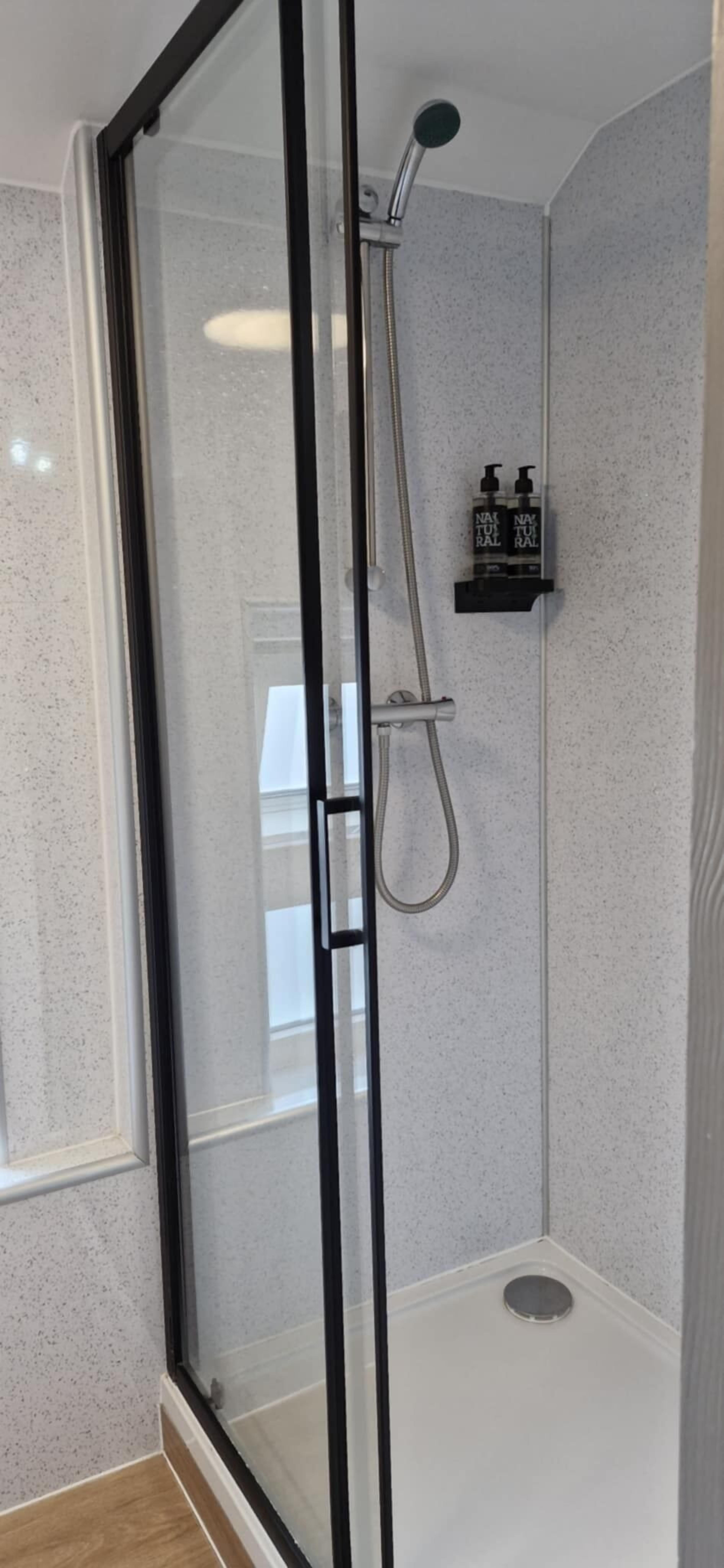 Une douche fermée par des parois en verre, dotée d’un pommeau de douche à main et d’un robinet fixé au mur.