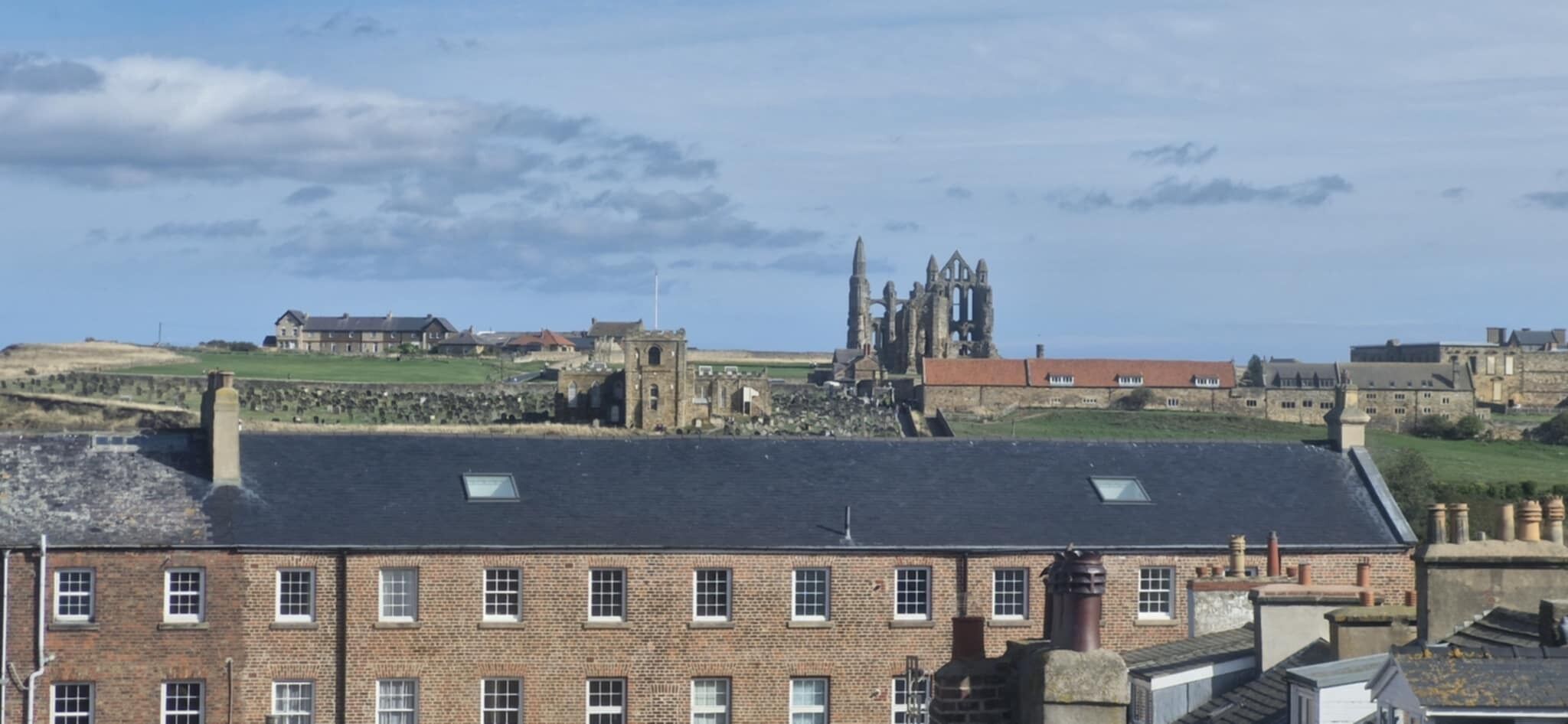A historic building with a prominent tower and ruins, surrounded by modern buildings and a grassy hill.