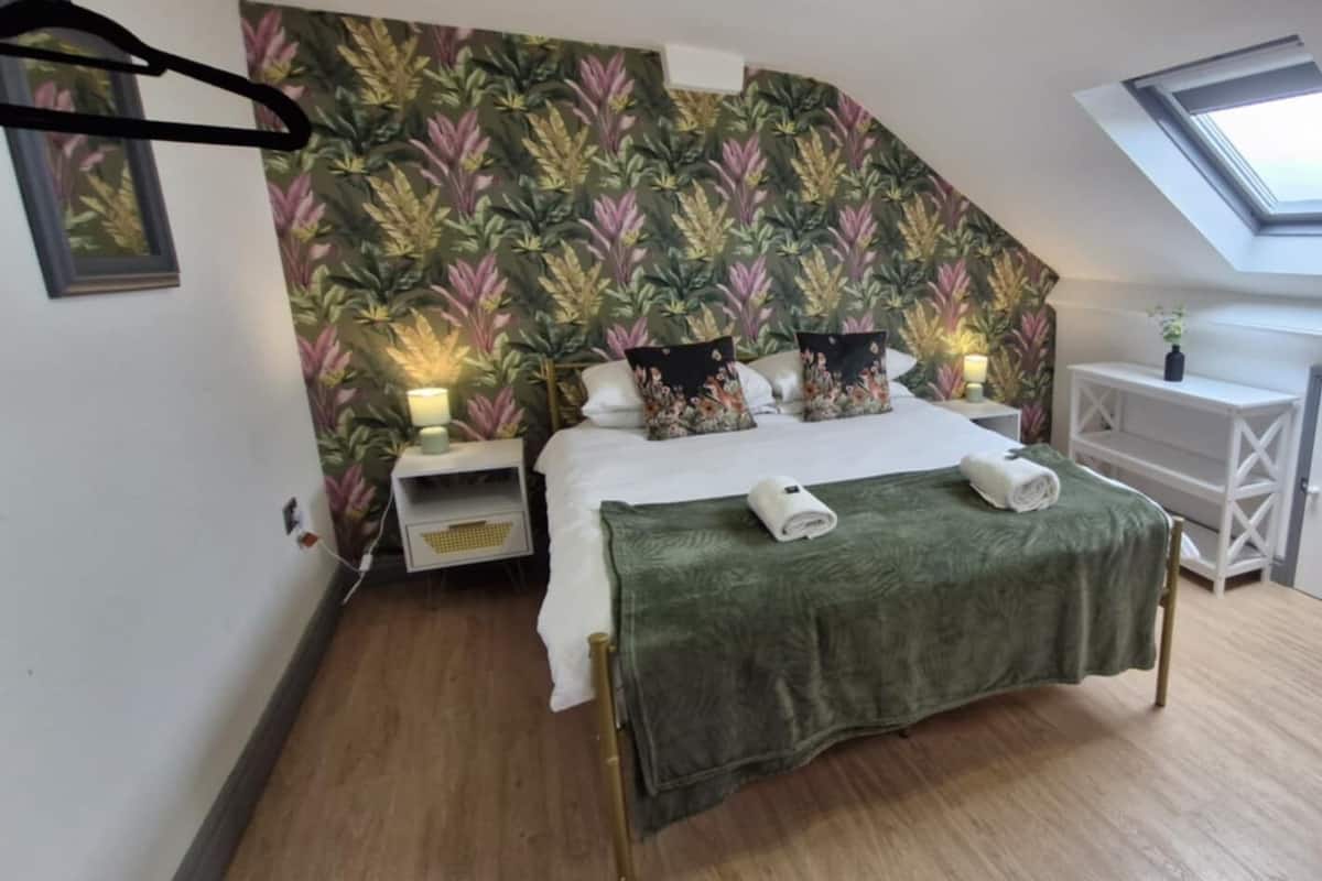 A bedroom with a sloped ceiling, a skylight, a bed with a green blanket, and a bedside table with a lamp.