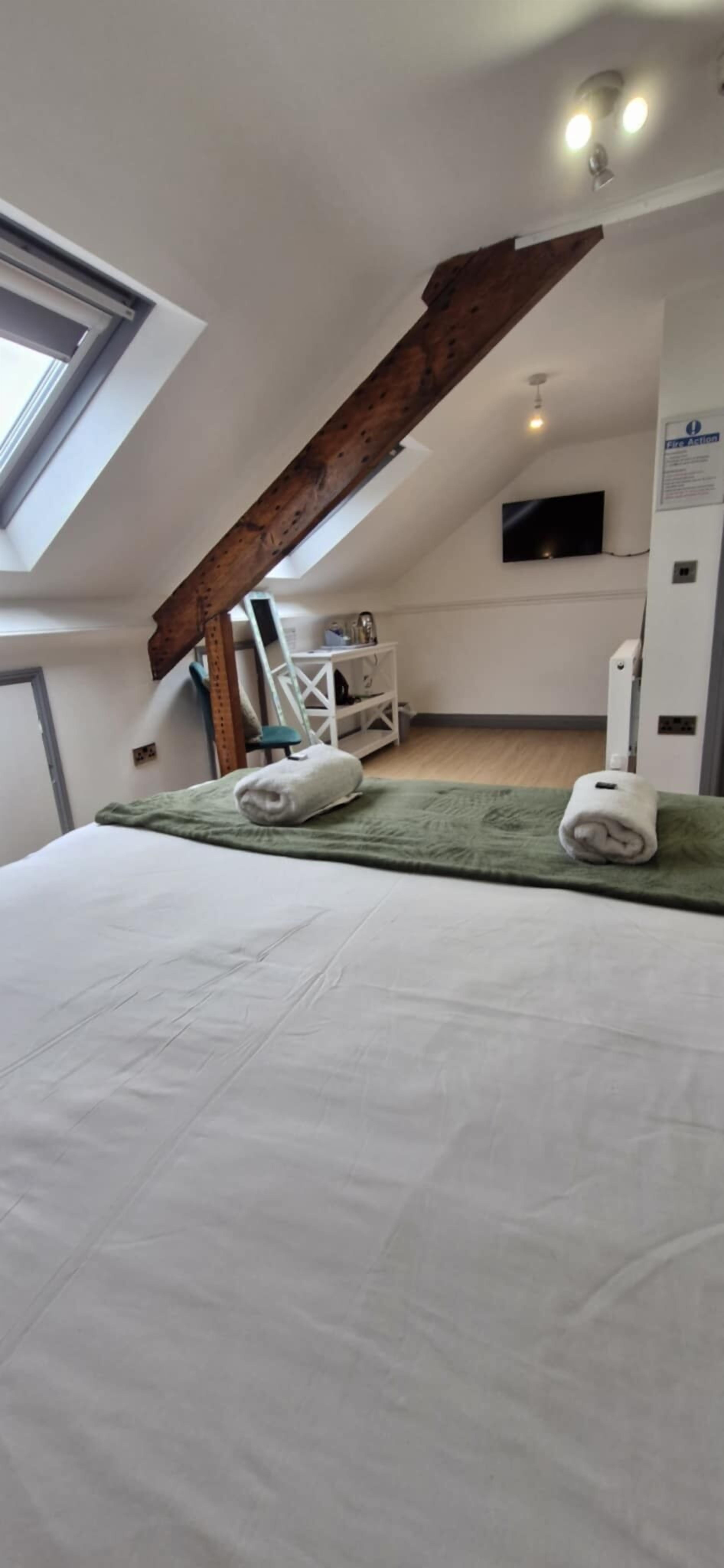 Un dormitorio con techo inclinado, tragaluces, cama con ropa de cama blanca, dos toallas enrolladas, televisor montado en la pared y un armario blanco.