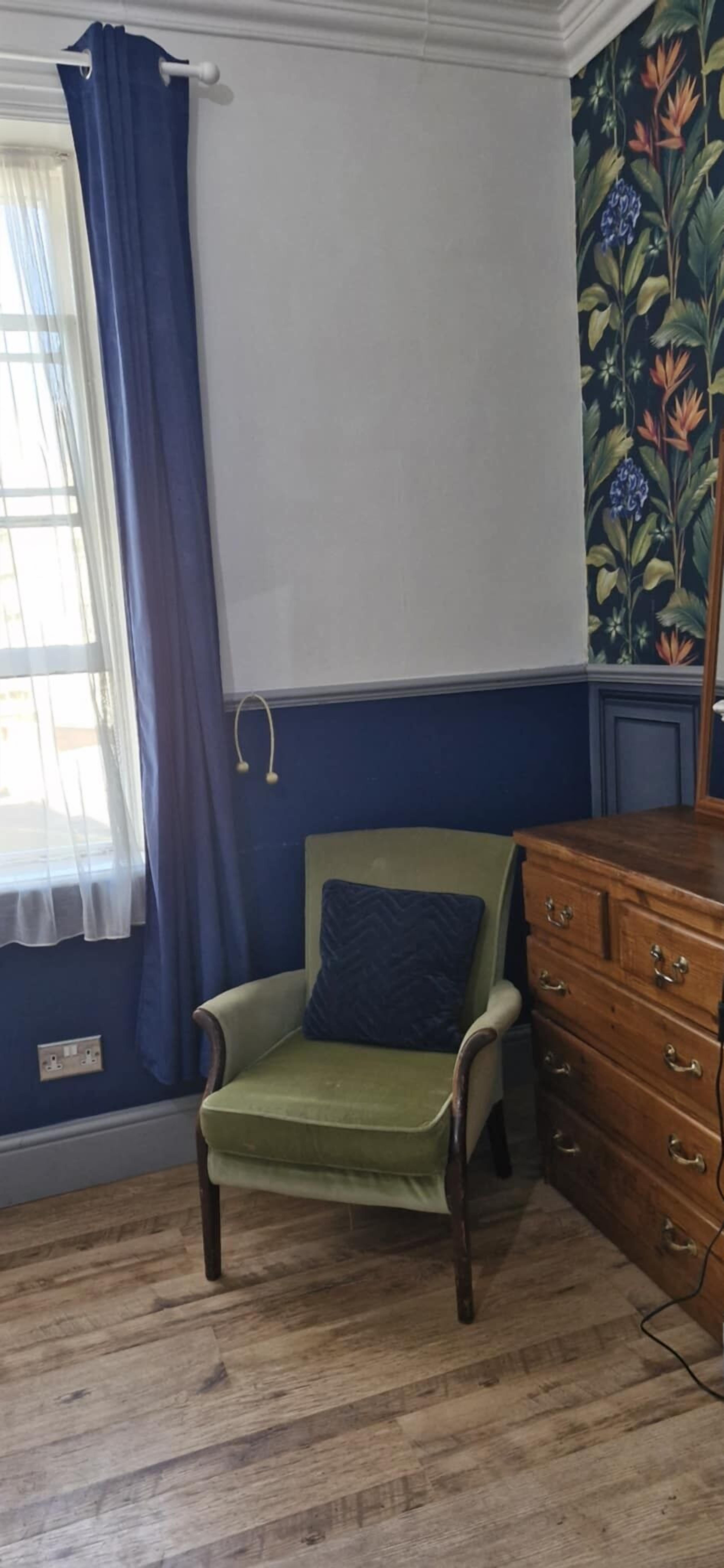 A room with a green armchair, a wooden dresser, a window with blue curtains, and a floral wallpaper.