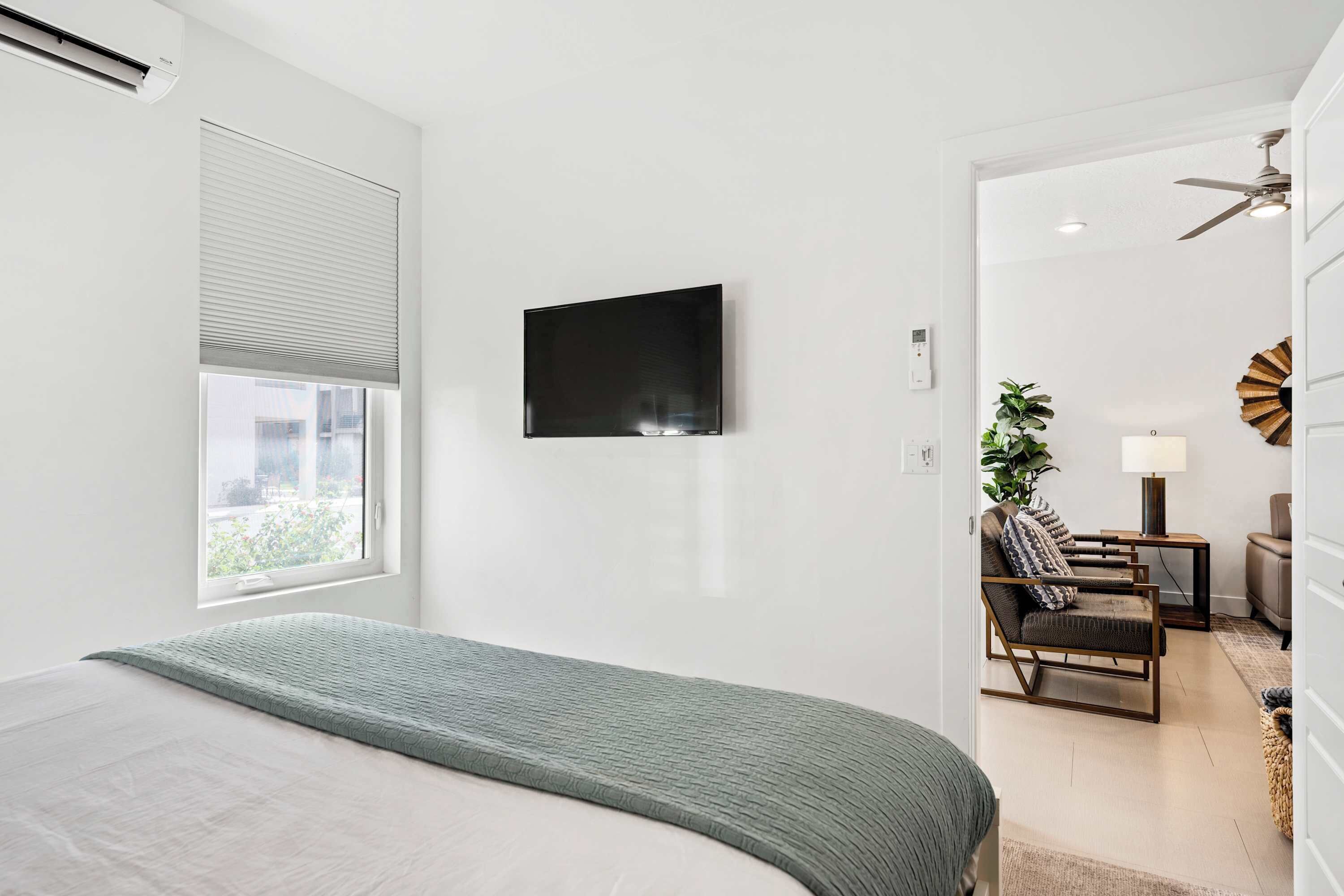 A modern bedroom with a large bed, a TV mounted on the wall, and a view into a living area with a sofa and a plant.