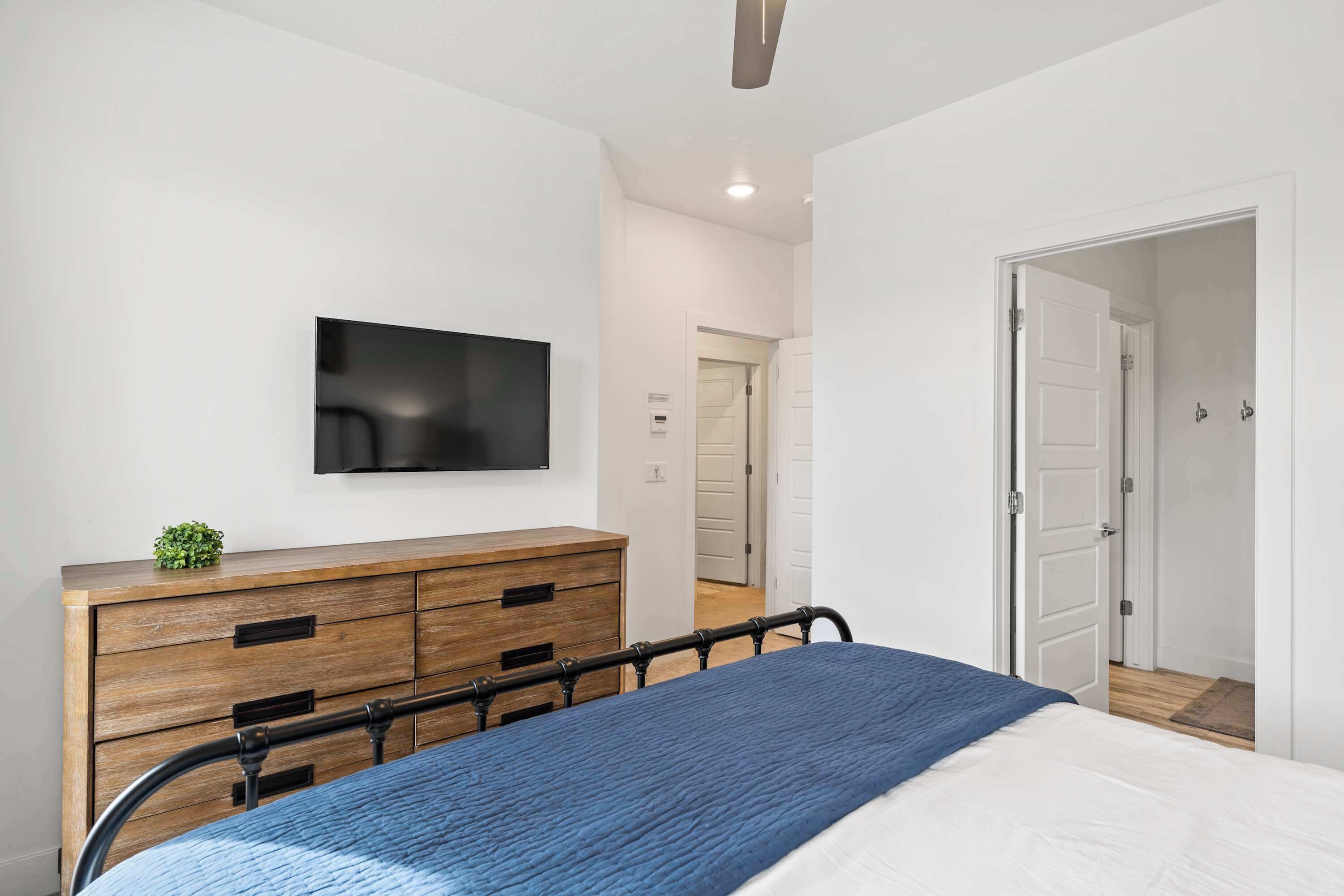 Um quarto com cama, cómoda de madeira, televisão fixada na parede e porta que dá para outro espaço.