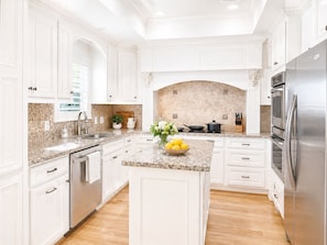 A modern kitchen with white cabinetry, granite countertops, a stainless steel refrigerator, and a central island with a bowl of fruit. - Cozy Waterfront I Fish Game Kayak I King 5BR House NASA (Houston)
