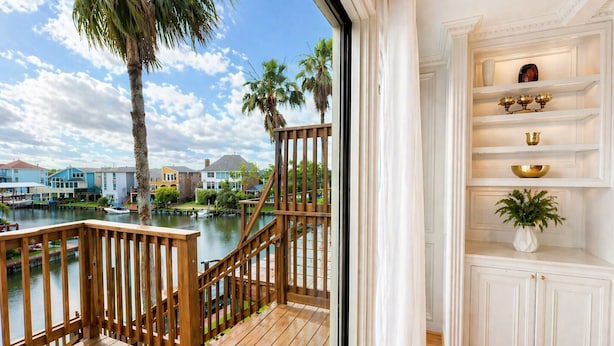 Terraza de madera con vistas a un puerto deportivo y casas, interior blanco con estantes y una planta.