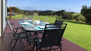 An outdoor dining area with a table set for four, surrounded by chairs, on a wooden deck with a view of a grassy landscape and hills in the background.
