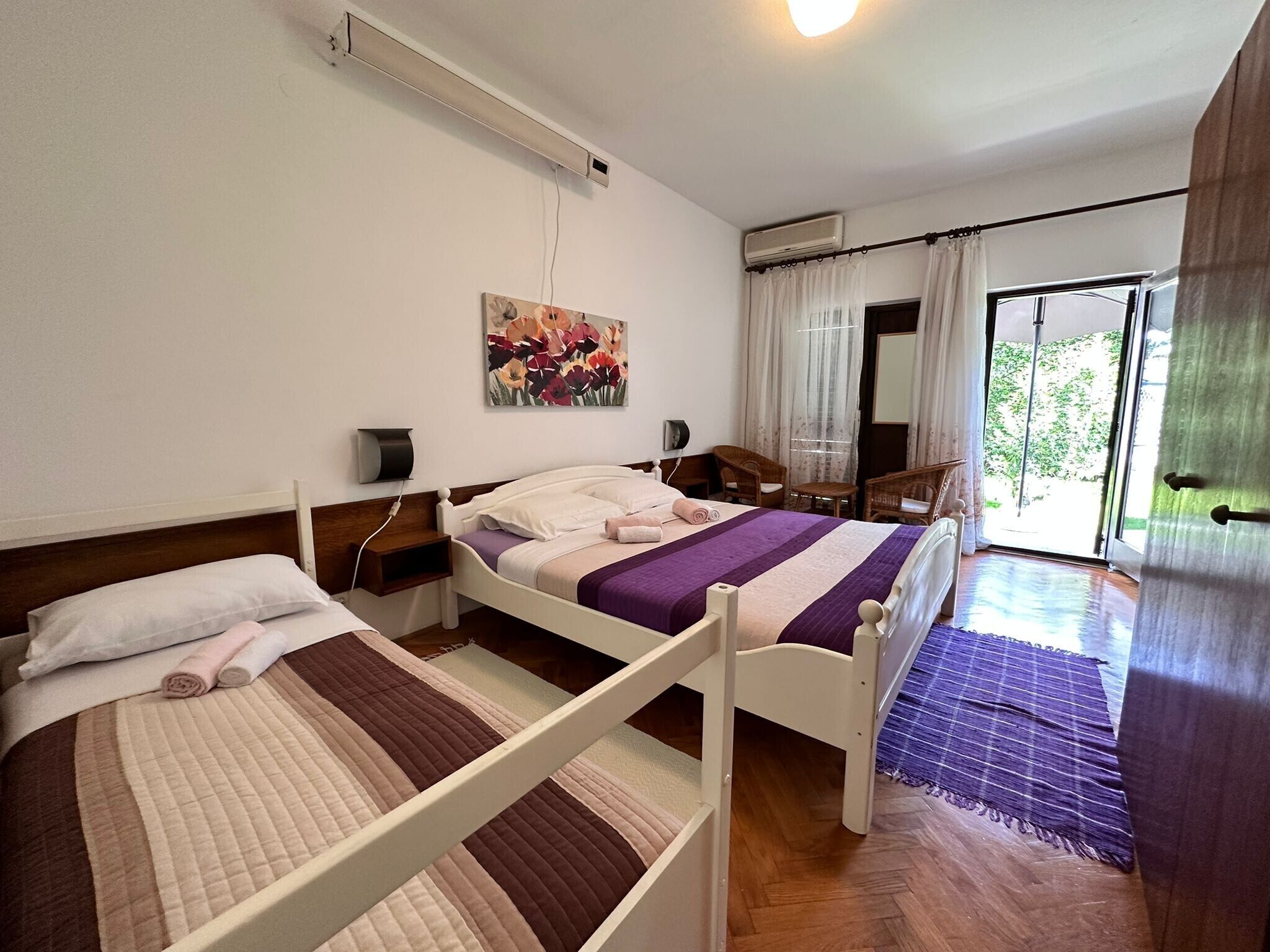 A bedroom with two beds, a painting, an air conditioning unit, a window with curtains, and a view of greenery outside.