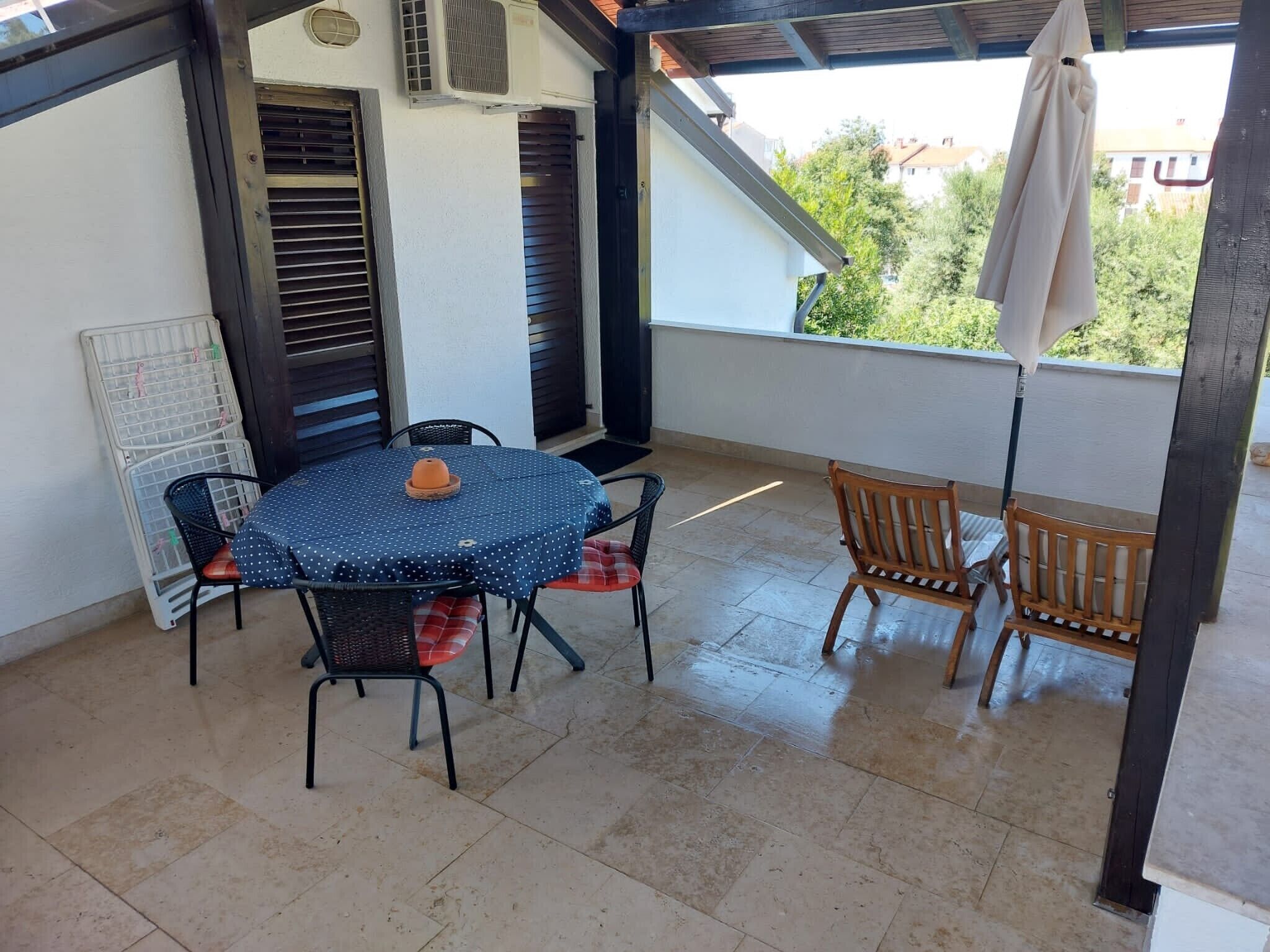 A covered patio with a round table and chairs, a wooden bench, and a view of greenery outside.