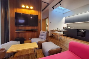 Een moderne woonkamer met een flatscreen televisie, houten lambrisering, een eethoek en een keuken met donkere kastjes.