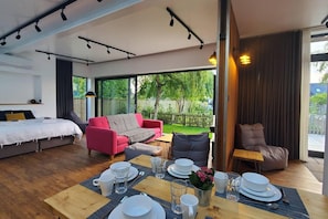 Een moderne hotelkamer met een groot bed, een rode bank, een zithoek met een tafel gedekt voor twee personen en uitzicht op de tuin door grote ramen.