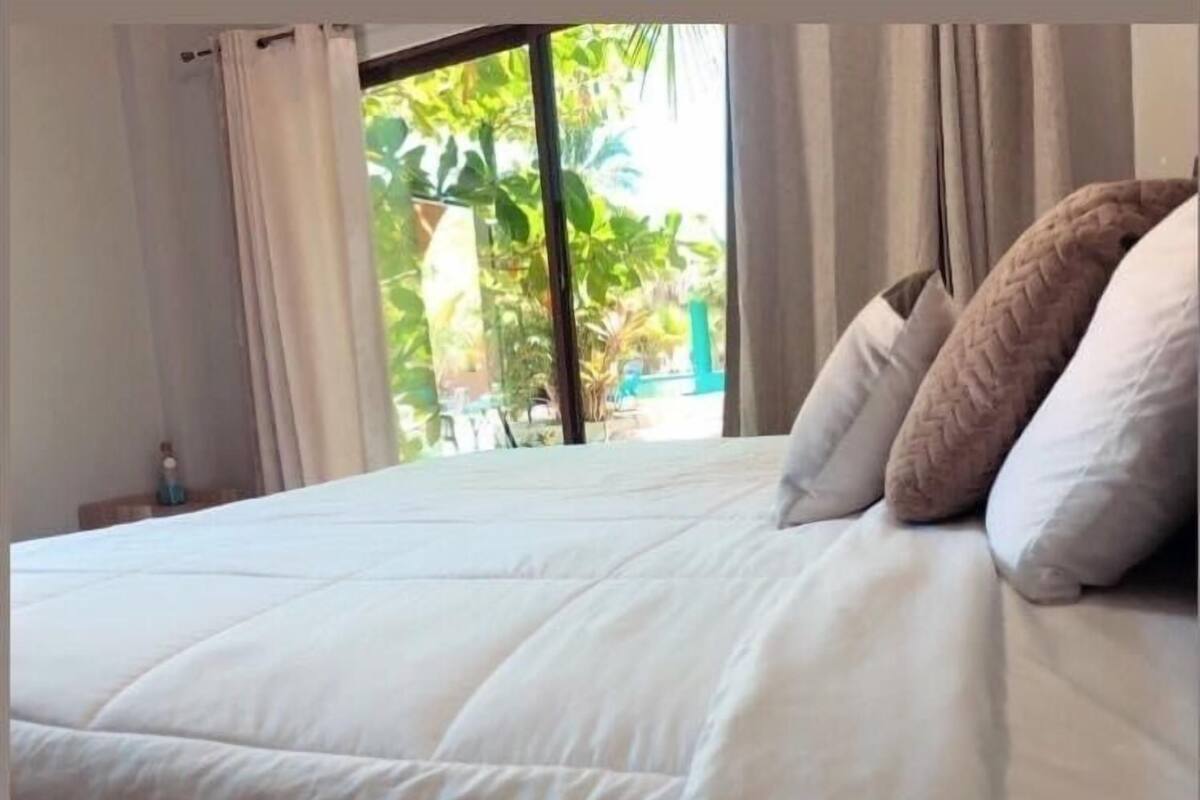 A neatly made bed with pillows, a window with curtains, and a view of greenery outside.