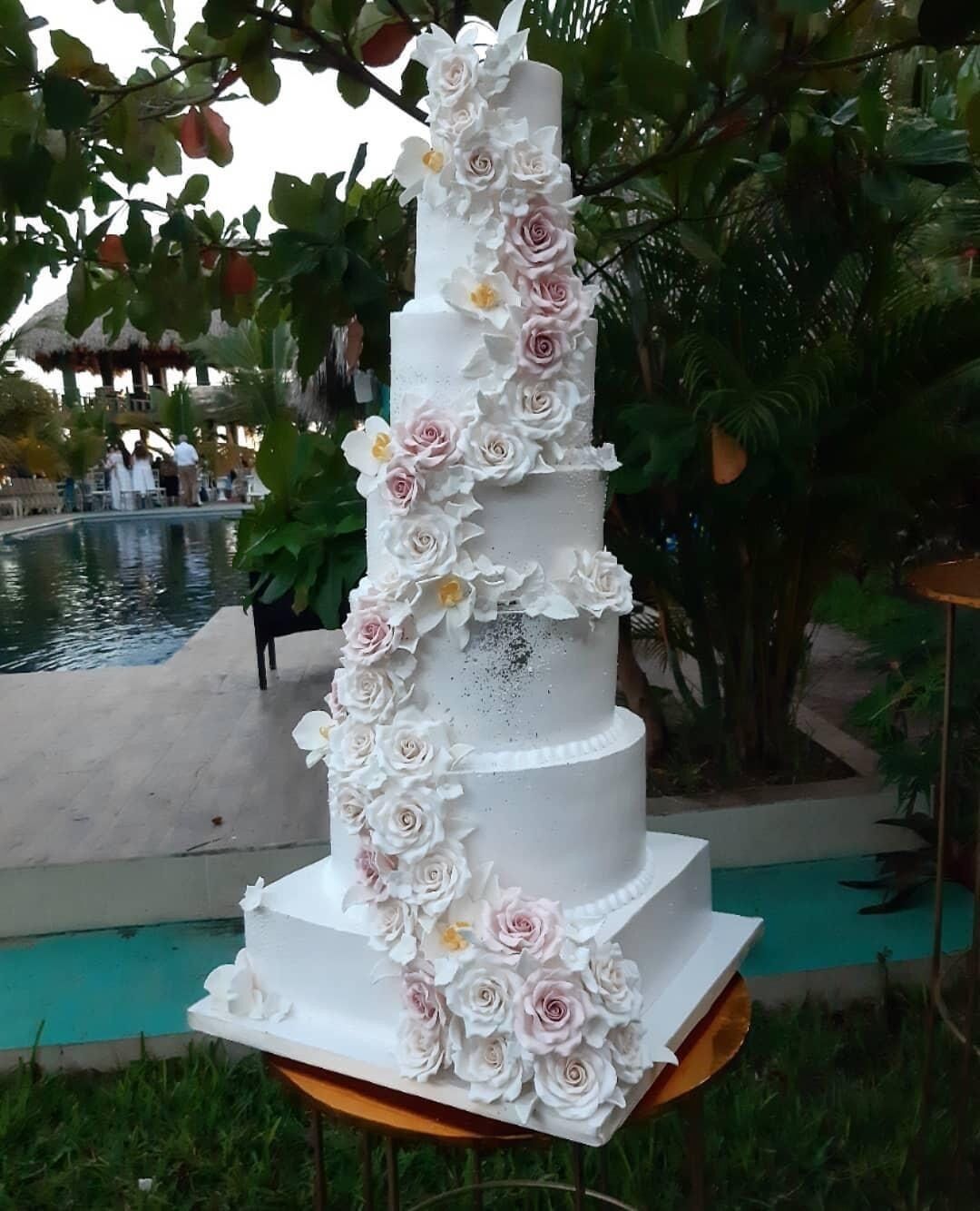 Un gâteau de mariage à plusieurs étages, décoré de fleurs blanches et roses, placé à l’extérieur, près d’une piscine.