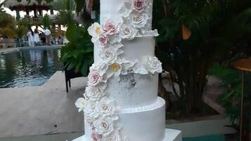 Un gâteau de mariage à plusieurs étages, décoré de fleurs blanches et roses, placé à l’extérieur, près d’une piscine.