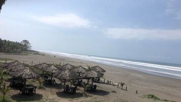 Zona de playa con sombrillas de paja y mesas, con vista al océano.