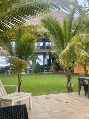A patio area with a wooden deck, a white lounge chair, and a black chair. There are palm trees and a building with a balcony in the background. - Sambo Mambo Beach Hotel (El Cuco)
