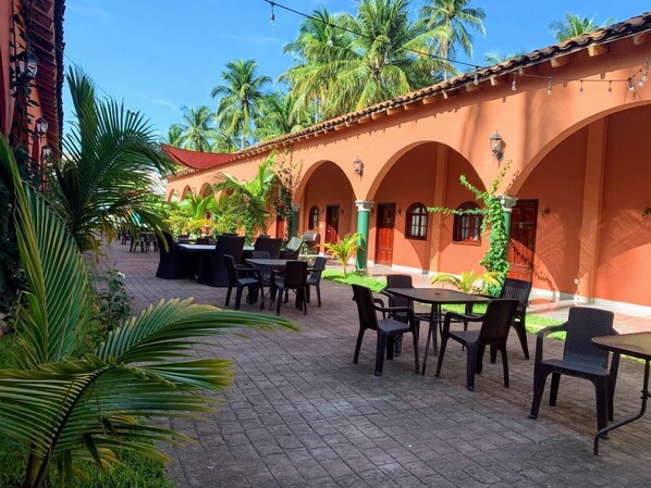 An outdoor patio area with tables and chairs, surrounded by palm trees and a building with arches. - Sambo Mambo Beach Hotel (El Cuco)