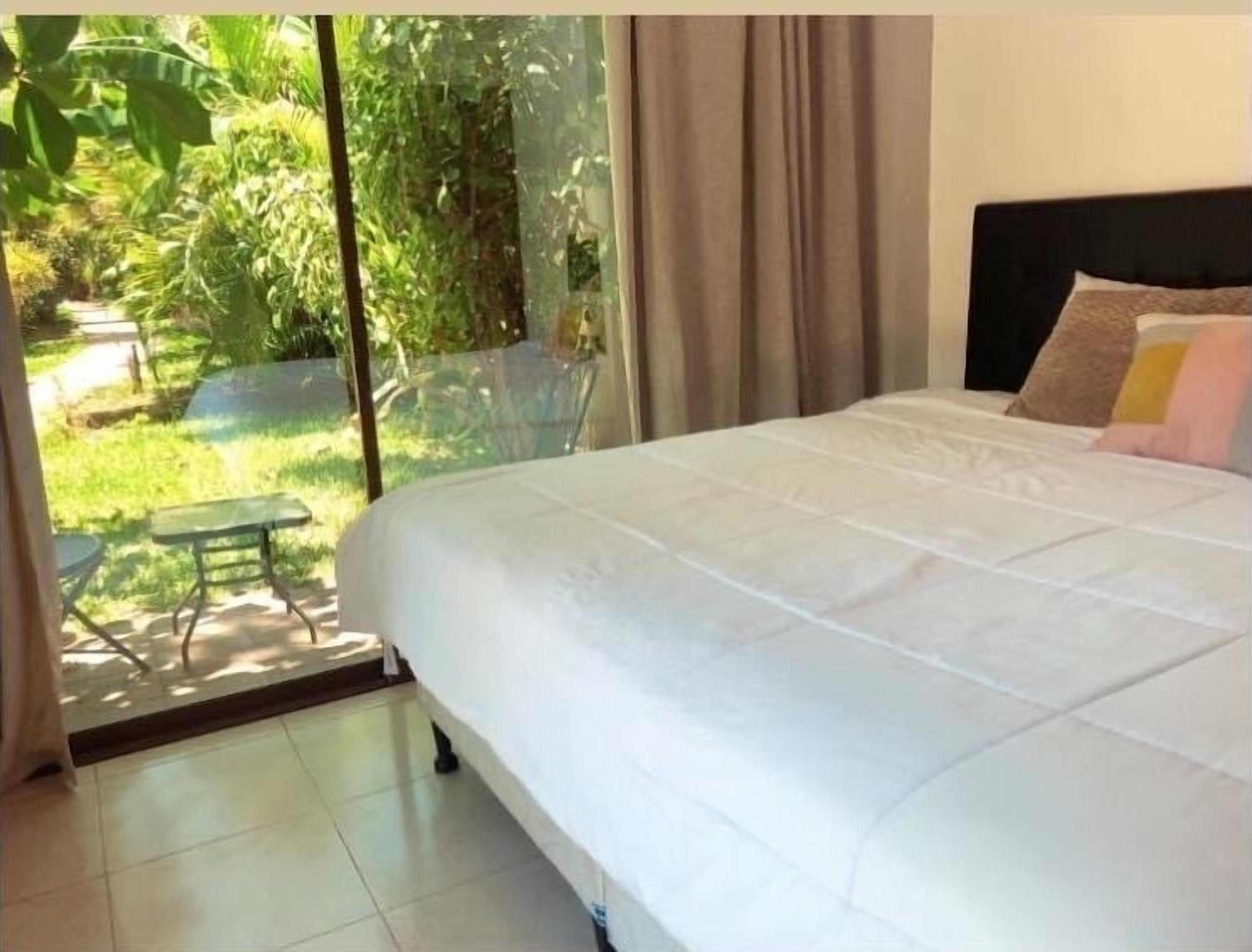 A bedroom with a large bed, a view of greenery outside, and a small table and chair by the window.