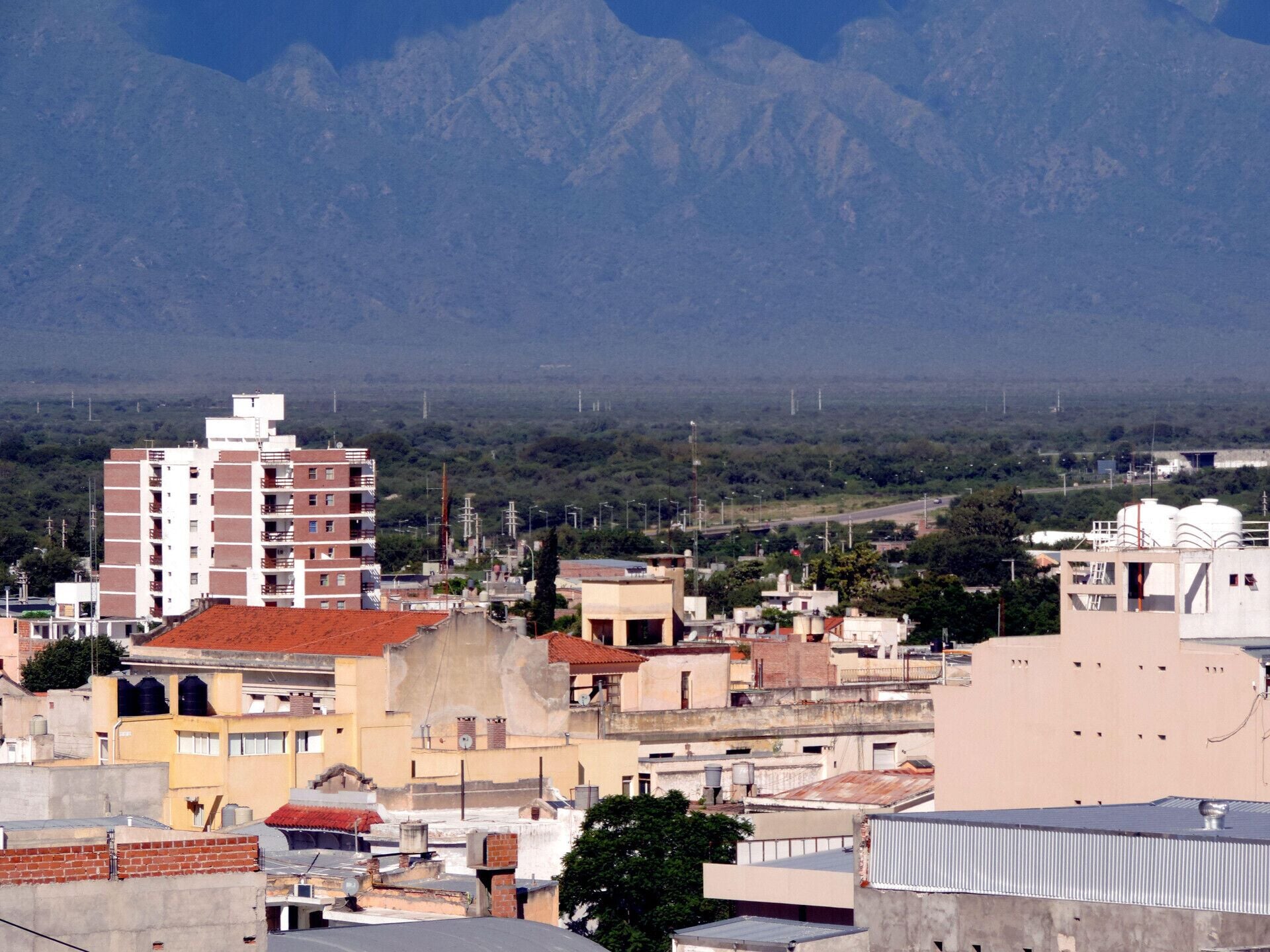 Paisagem urbana com edifícios, incluindo uma estrutura vermelha e branca em destaque, e uma cadeia de montanhas ao fundo.