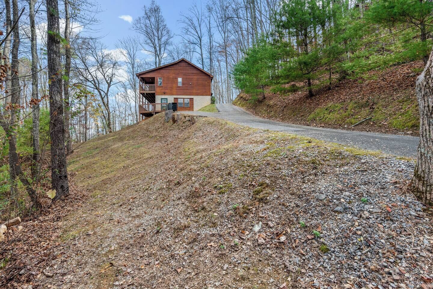 A cabin with a balcony, surrounded by a forested area and a gravel path leading to a road.