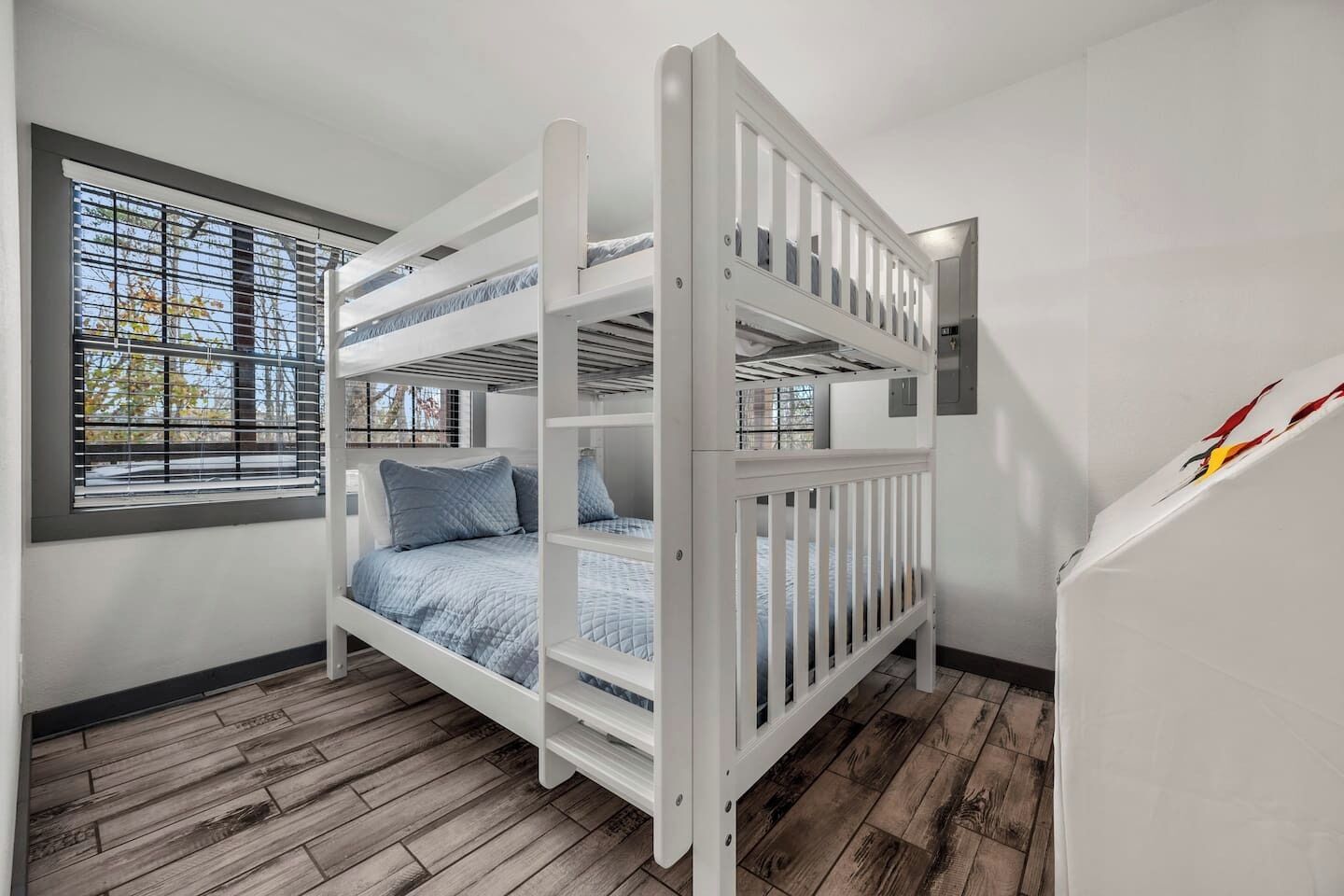A bunk bed with a white frame and blue bedding, a window with blinds, and a wooden floor.