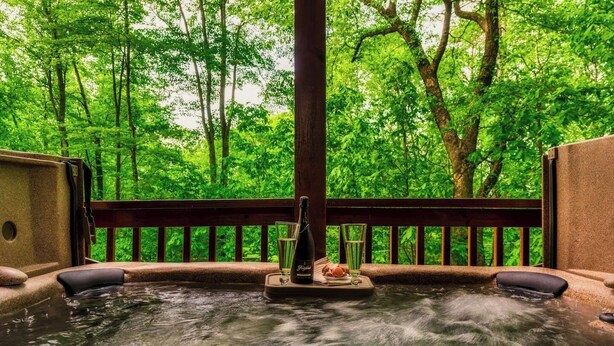 Un bain thermal en plein air, entouré d’une végétation luxuriante, avec une terrasse en bois et une bouteille de champagne.