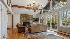 A spacious living room with a stone fireplace, wooden beams, and large windows offering a view of autumn foliage. - Spacious Stratton Mountain Home  - Perfect for families! (Winhall)