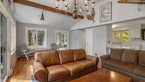 A spacious living room with a brown leather sofa, a wooden coffee table, and a chandelier. There are large windows and a ski rack on the wall. - Spacious Stratton Mountain Home  - Perfect for families! (Winhall)