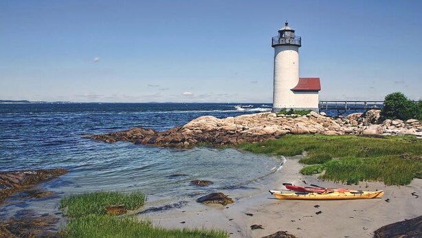 Un faro en una costa rocosa con aguas tranquilas, un kayak en la arena y un cielo despejado.