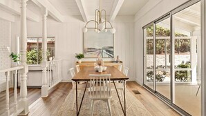 Uma sala de jantar com mesa de madeira, cadeiras brancas, um candelabro e uma grande janela com vista para o verde.