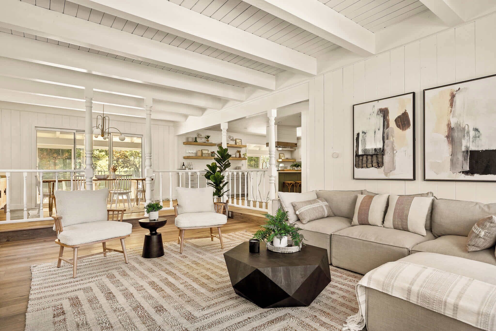 A spacious living room with a large sofa, two armchairs, a coffee table, and a dining area with a table and chairs. There are two large windows providing natural light and a view of the outdoors.