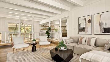 A spacious living room with a large sofa, two armchairs, a coffee table, and a dining area with a table and chairs. There are two large windows providing natural light and a view of the outdoors.