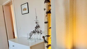 Ein Zimmer mit weißem Schminktisch, einem Birkenstamm, einem gerahmten Bild an der Wand und einer beleuchteten Lampe.