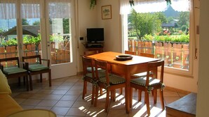 Ein Essbereich mit Holztisch und Stühlen, einem Fernseher auf einem Schrank und großen Fenstern mit Vorhängen.