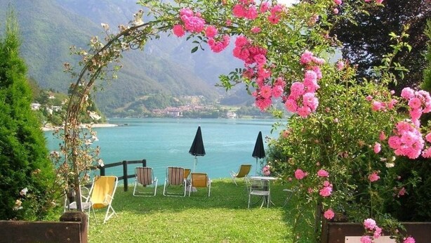 Ein Garten mit Seeblick und Bergblick, mit Sitzgelegenheiten im Freien und einer Pergola mit rosa Blüten.