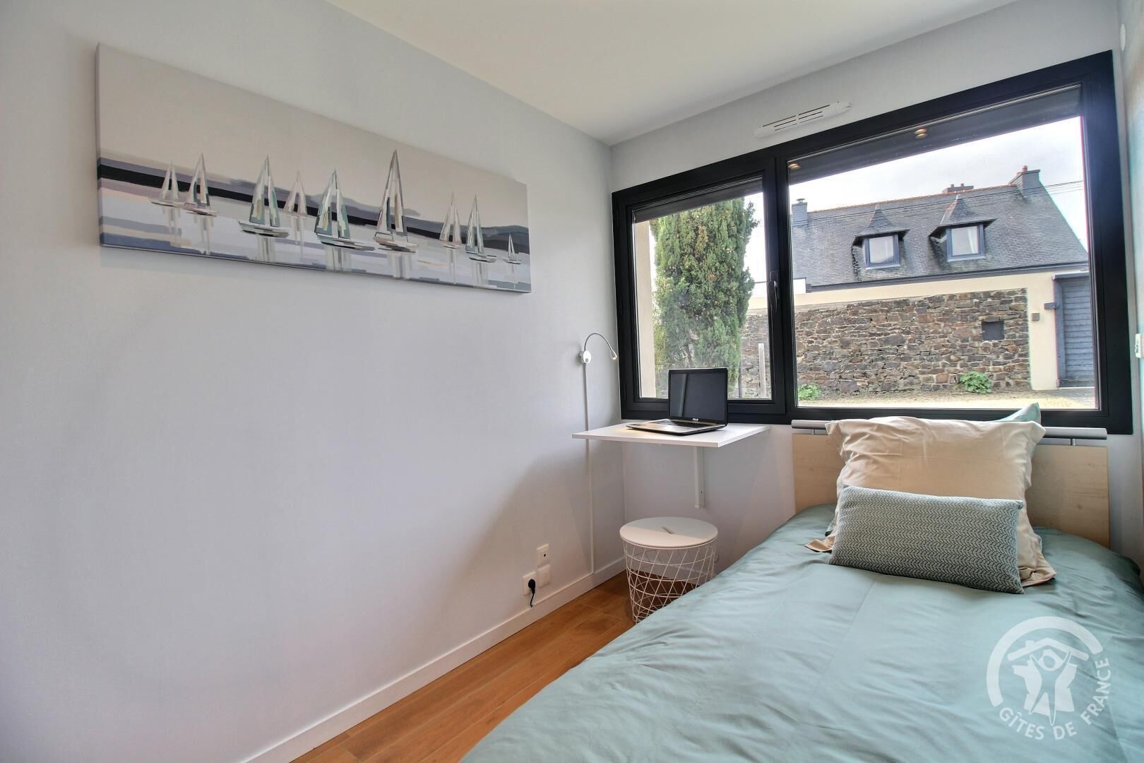 A bedroom with a bed, a laptop on a window sill, a wall-mounted artwork of sailboats, and a view of a stone building outside.