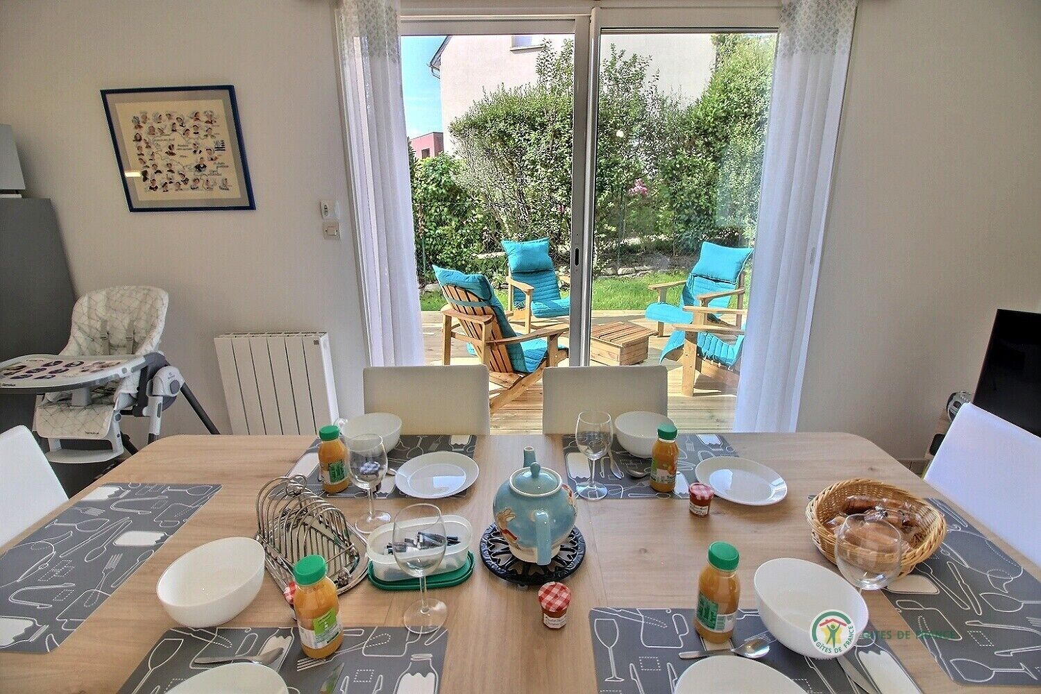 A dining area with a table set for two, a high chair, and a view of the garden through glass doors.