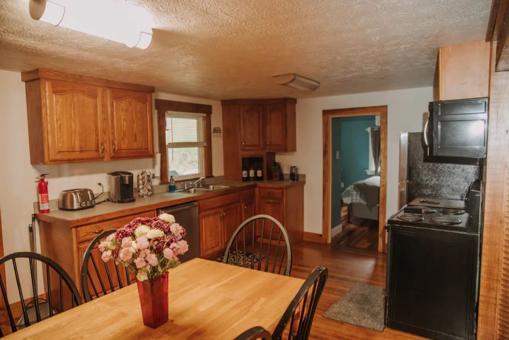 A compact kitchen with wooden cabinets, a dining table with chairs, and a black stove.