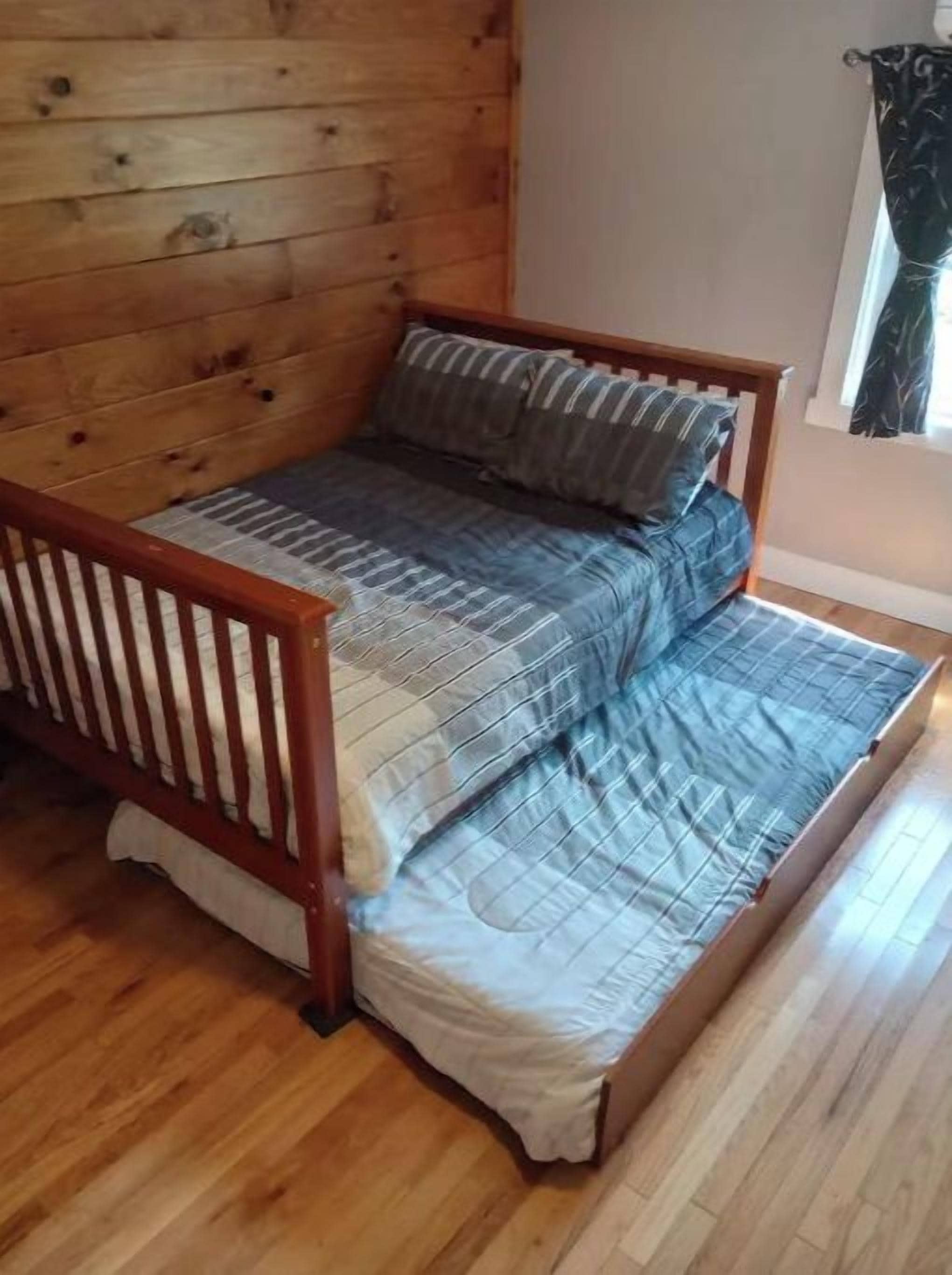 A wooden bed with a mattress and pillows in a room with wooden flooring and a window with curtains.
