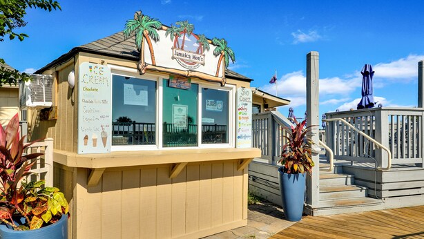 Un puesto pequeño de helados con un cartel de menú, situado en una terraza de madera con barandilla y plantas en macetas.