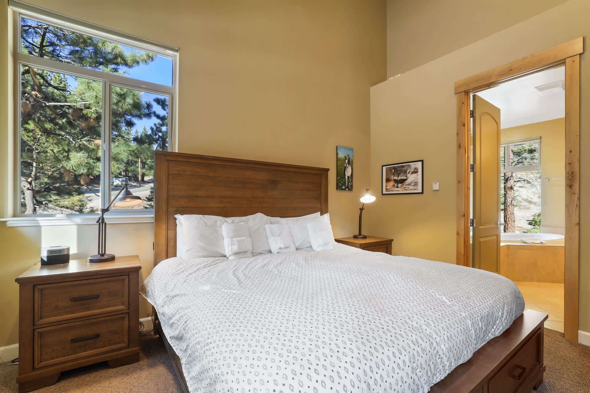 A bedroom with a wooden headboard, a large bed with white bedding, a nightstand with a lamp, and a window with a view of trees.