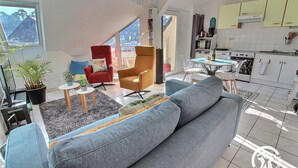 A modern living room with a grey sofa, red armchair, and a small coffee table. There is a kitchen area with white cabinets and a dining table. - Trestraou - Perros Guirec (PERROS GUIREC)