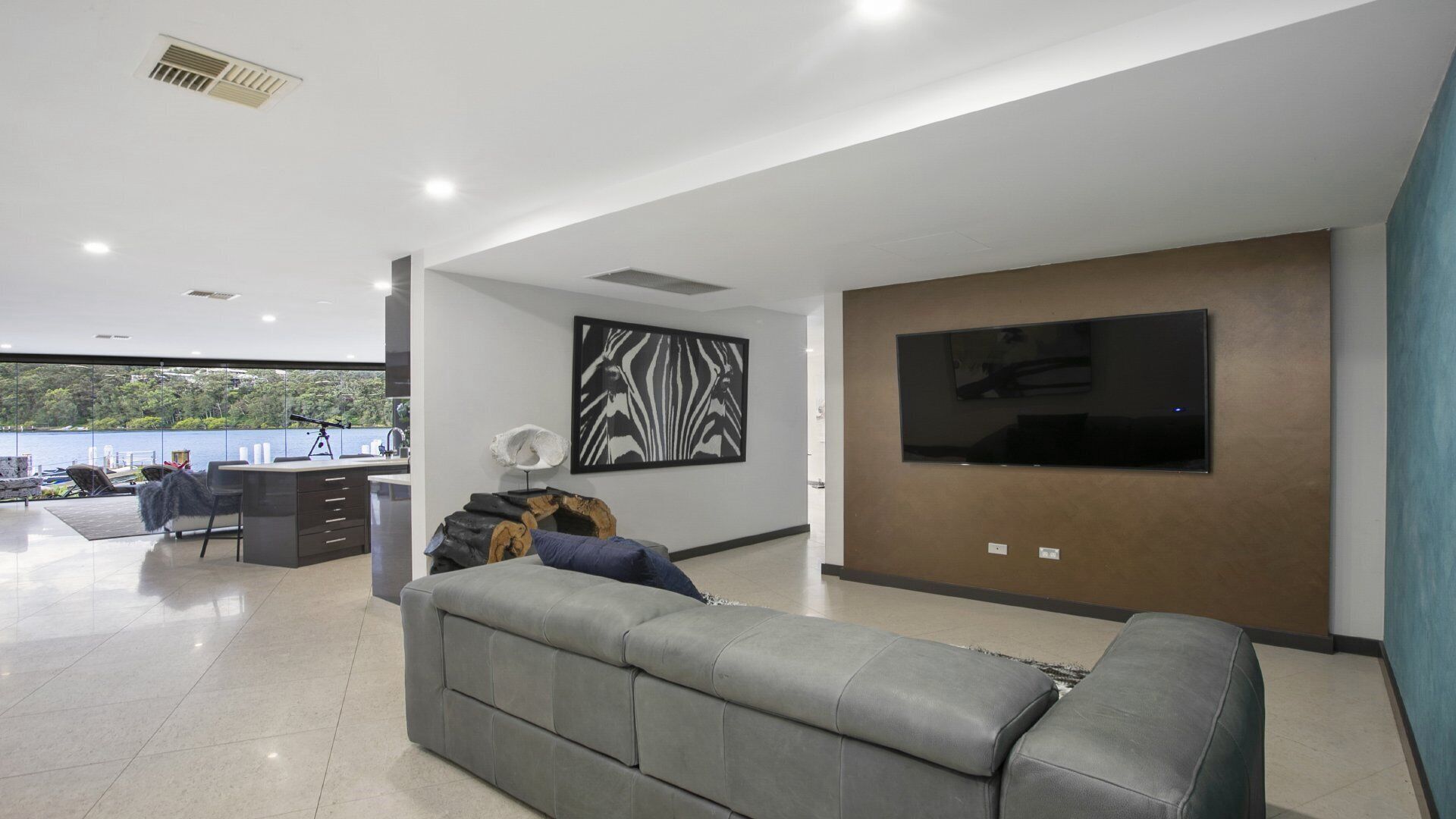 A modern living room with a grey sofa, a flat-screen TV mounted on a brown wall, and a large window overlooking a body of water.