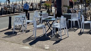 Café al aire libre con sillas y mesas blancas, sombrillas y vista a la playa y al mar.