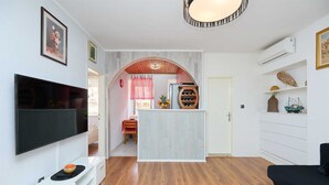 A compact living space with a flat-screen TV, a wooden dining table, a kitchen area with a wine rack, and a small island.